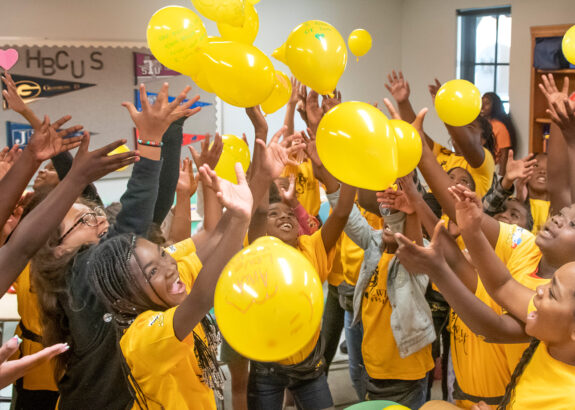 a group of girls wearing yellow tee shirts releases balloons inside a large room